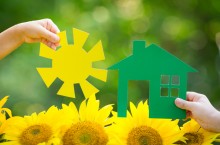 Two hands holding paper house and sun over flowers